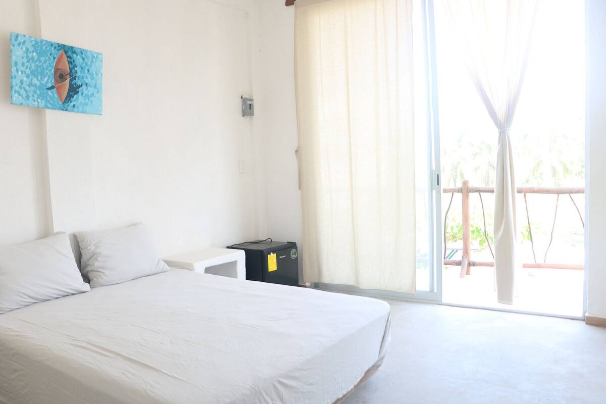 A bedroom with a bed, a small bedside table, a power outlet, and a window with curtains.