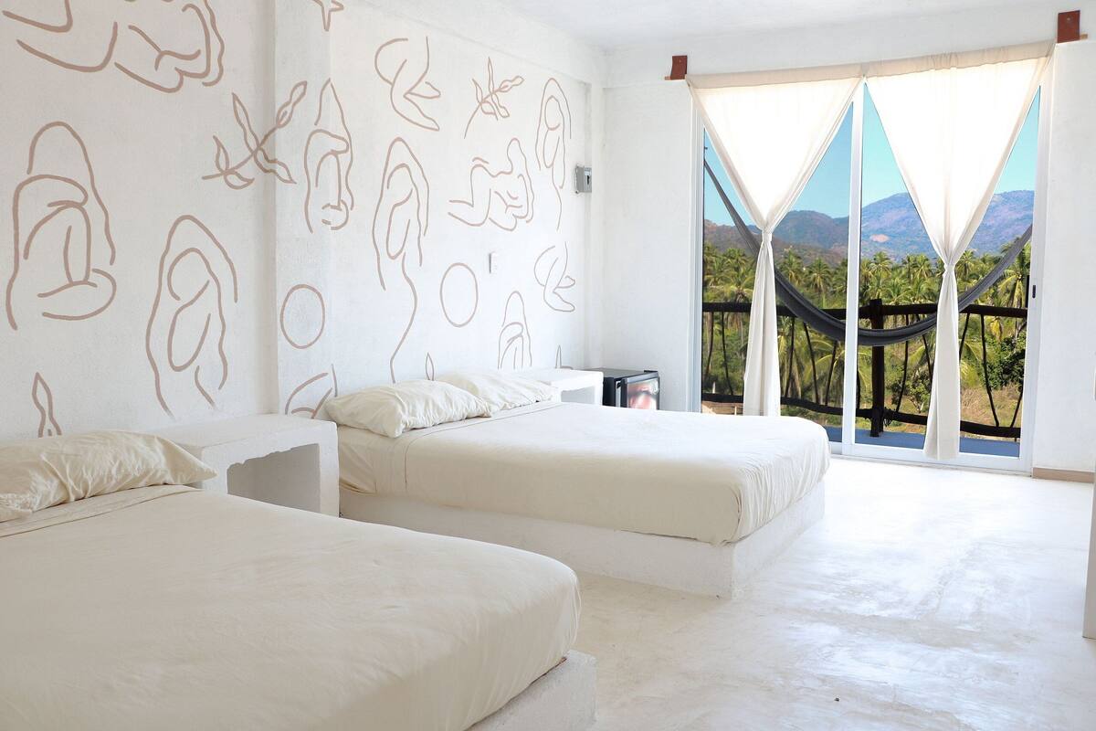 A bedroom with two beds, a large window with a view of palm trees and mountains, and a hammock outside.