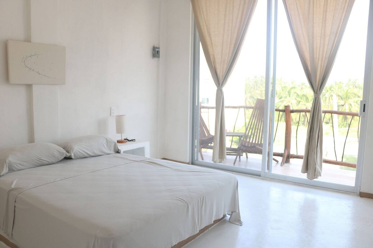 A bedroom with a large bed, a nightstand, a lamp, and a view of the outdoors through a sliding glass door.