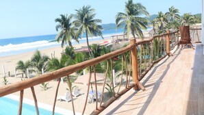 Un balcón con barandilla de madera que da a una playa con palmeras y una piscina.