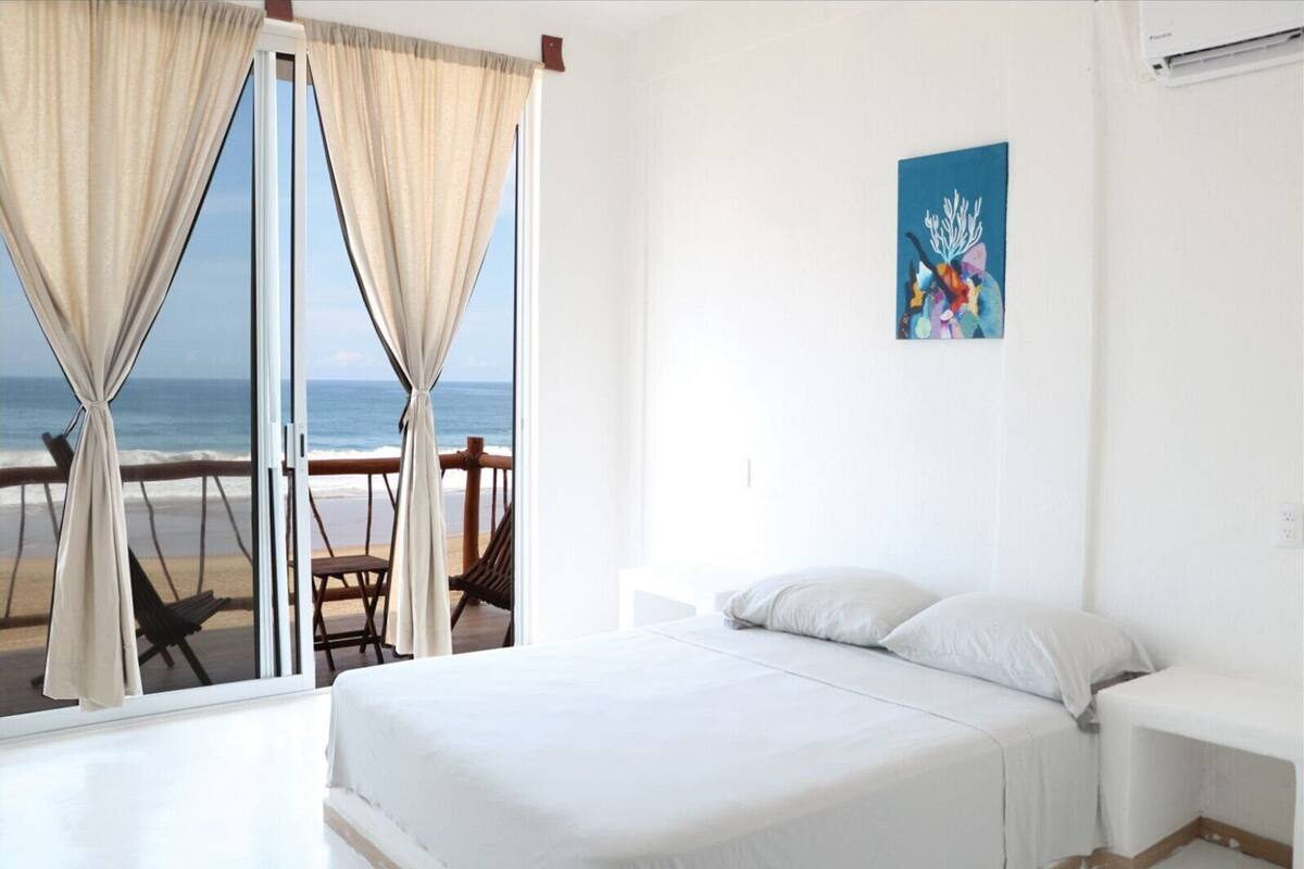 A hotel room with a bed, a nightstand, and a view of the beach through a sliding door.