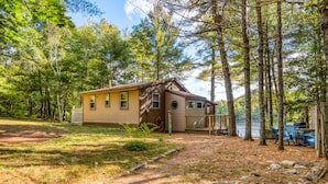 A cabin with a small deck and a grill area, surrounded by trees and a lake. - Lakefront Cottage with Private Dock  and Kayaks. Dog-Friendly (Newfield)