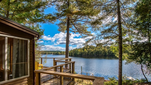 A wooden cabin with a deck overlooking a lake, surrounded by trees. - Lakefront Cottage with Private Dock  and Kayaks. Dog-Friendly (Newfield)