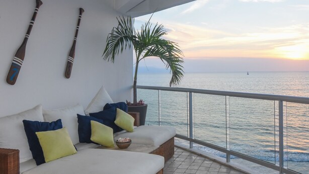 Un balcon avec un canapé blanc doté de coussins, une plante en pot et une vue sur la mer au coucher du soleil.
