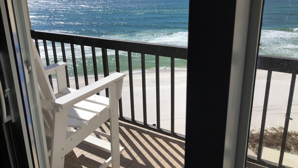 Un balcon avec une chaise blanche, donnant sur une plage où l’on peut voir des vagues et un ciel dégagé.