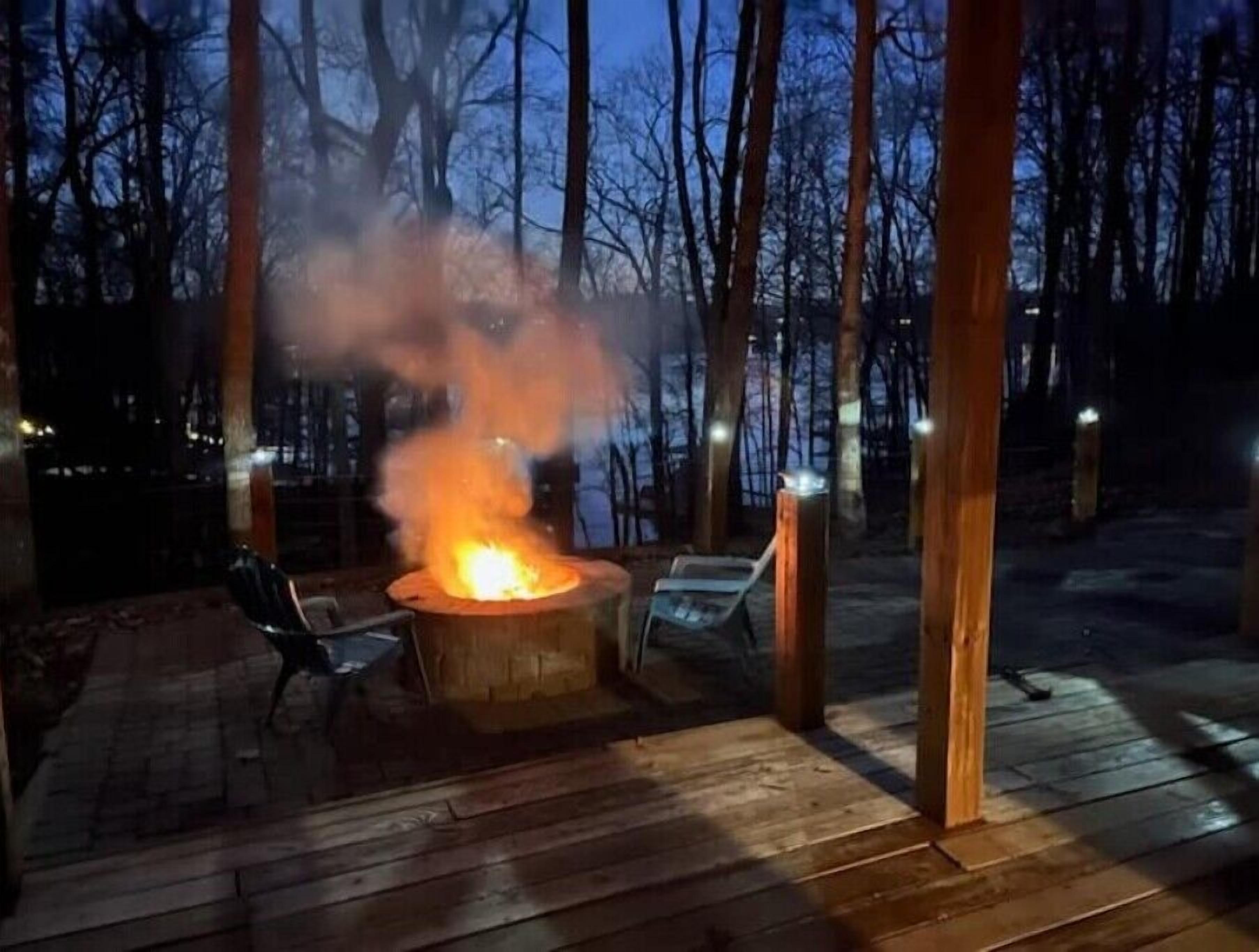 A fire pit in a wooded area with a wooden deck and chairs.