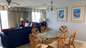 A living room with a dark blue sofa, a glass-top dining table surrounded by wooden chairs, a ceiling fan, and two framed artworks on the wall. - 3BR 3BA Oceanfront 7th Floor N. Myrtle Beach Condo (North Myrtle Beach)