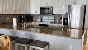 A modern kitchen with white cabinets, a granite countertop, a microwave, a stove, and a refrigerator. There are three wooden stools with black cushions. - 3BR 3BA Oceanfront 7th Floor N. Myrtle Beach Condo (North Myrtle Beach)