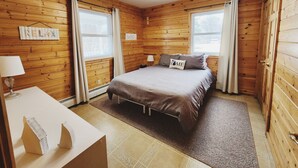 Ein gemütliches Schlafzimmer mit Holzwänden, einem Bett mit einem 'HOME'-Kissen, einem Nachttisch mit Lampe und einer kleinen Häuser-Dekoration.