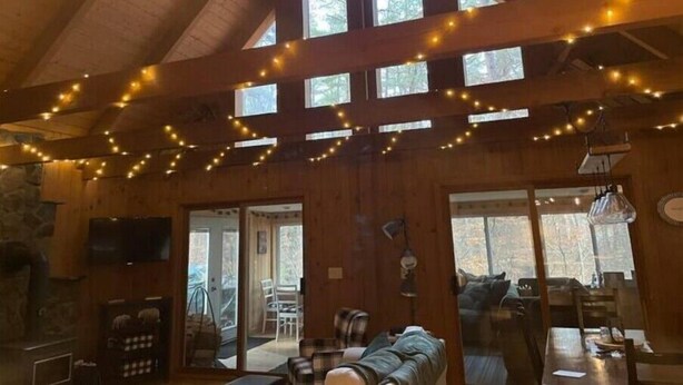 Interior acogedor de una cabaña con techo abovedado, luces de adorno, chimenea y un ventanal con vistas a un bosque.