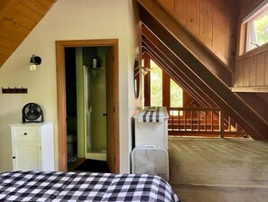 A bedroom with a checkered bedspread, a white dresser, a fan, and a view of a wooden staircase leading to a balcony.