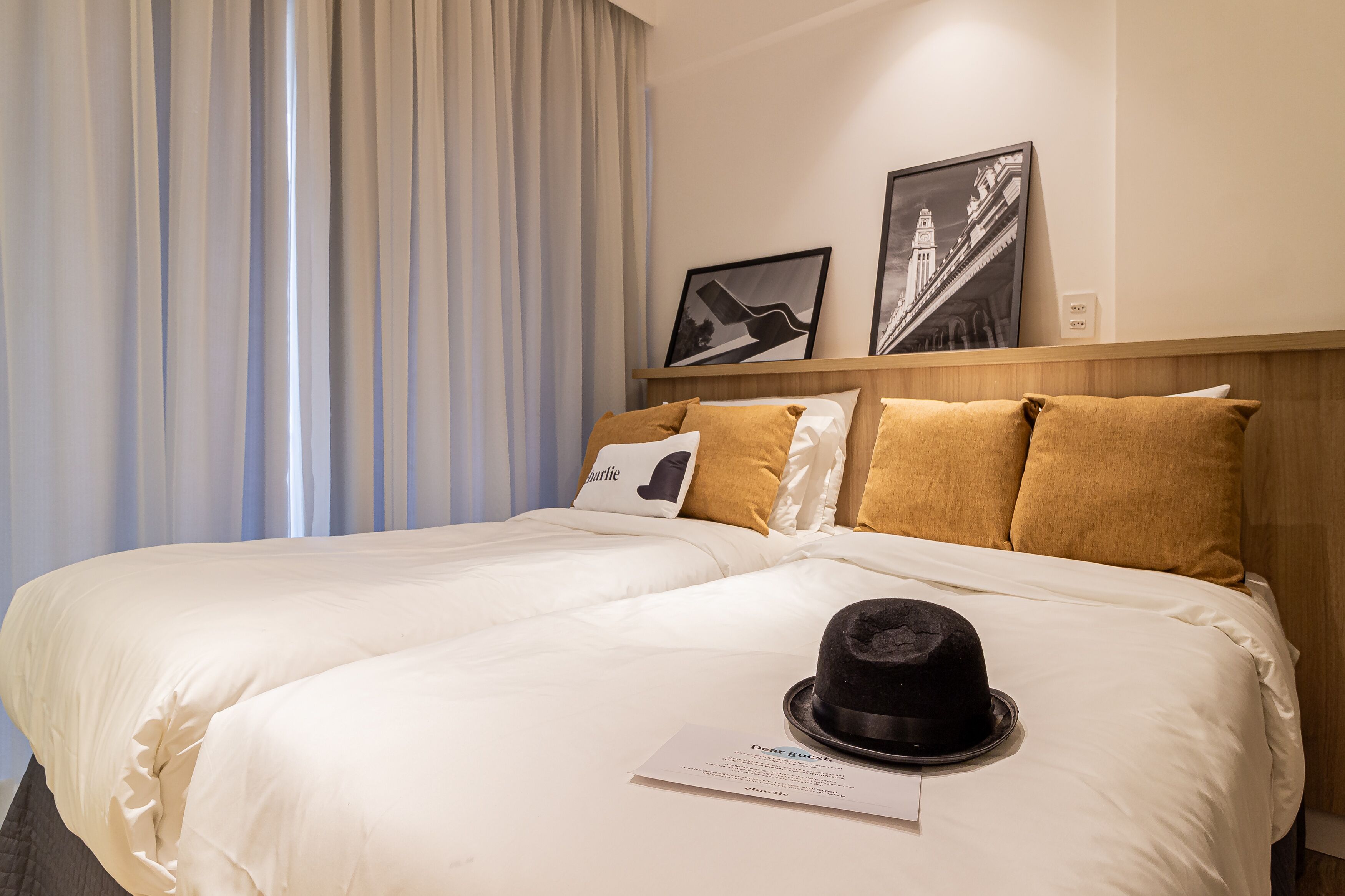A hotel room with two beds, a black hat on the bed, and two framed pictures on the wall.