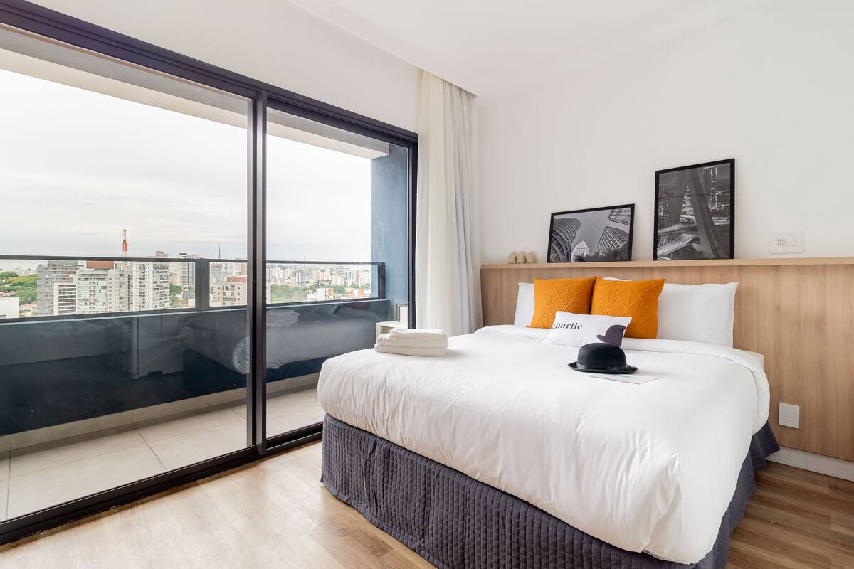 Ein Hotelzimmer mit einem großen Bett, Blick auf die Stadt durch eine Schiebetür und zwei gerahmte Bilder an der Wand.