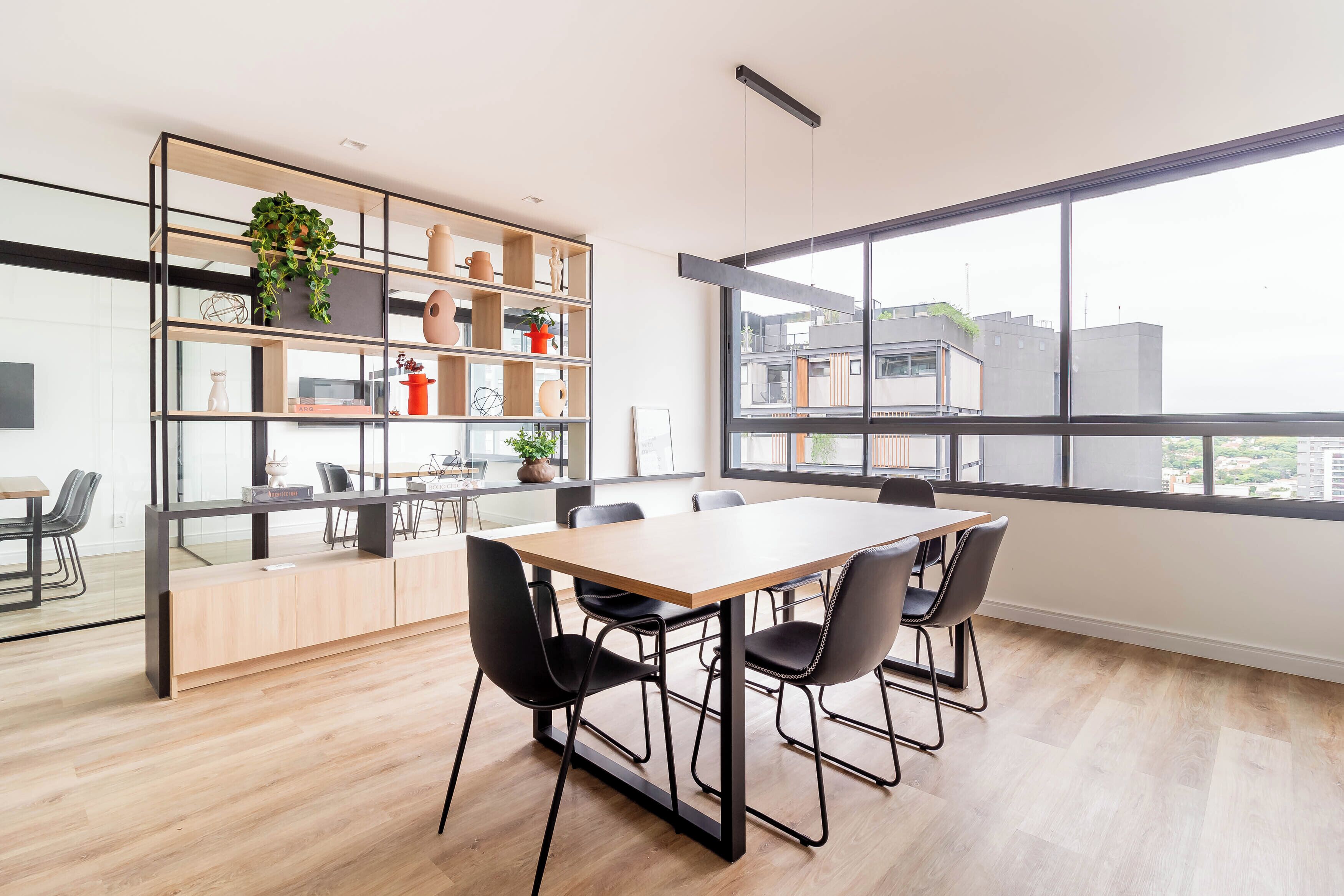 Een moderne vergaderruimte met een grote tafel, vier stoelen, een glazen scheidingswand en een houten wandplank met decoratieve objecten.