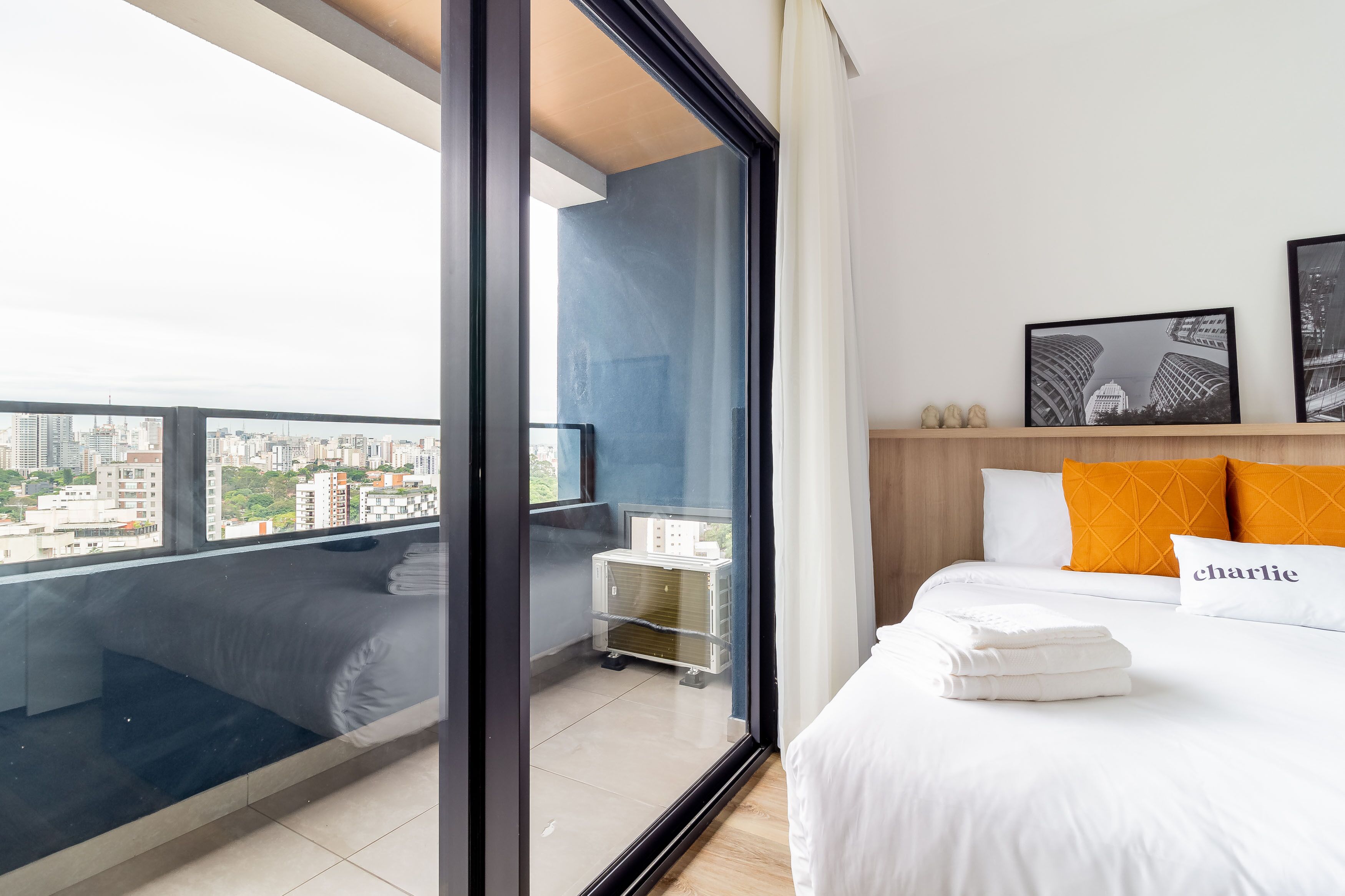 Una habitación de hotel moderna con un ventanal grande, una cama con ropa blanca, una almohada 'charlie' y cuadros enmarcados en la pared.