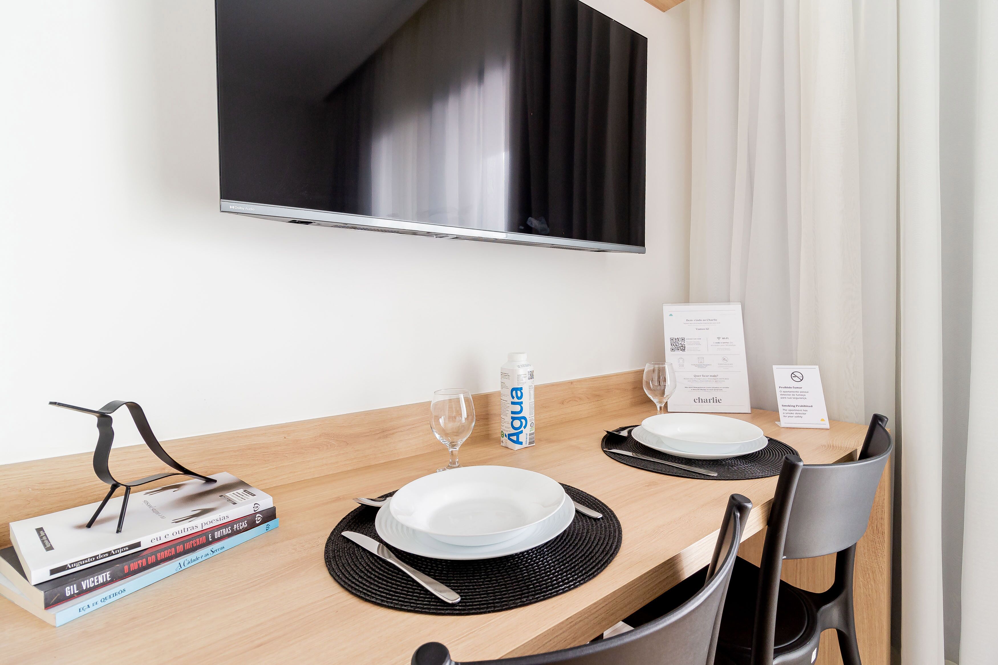 A hotel room with a wooden dining table set for two, a flat-screen TV mounted on the wall, a minibar, and a window with curtains.