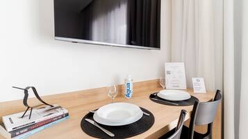Habitación de hotel con mesa de comedor de madera dispuesta para dos personas, un televisor de pantalla plana en la pared, un minibar y una ventana con cortinas.