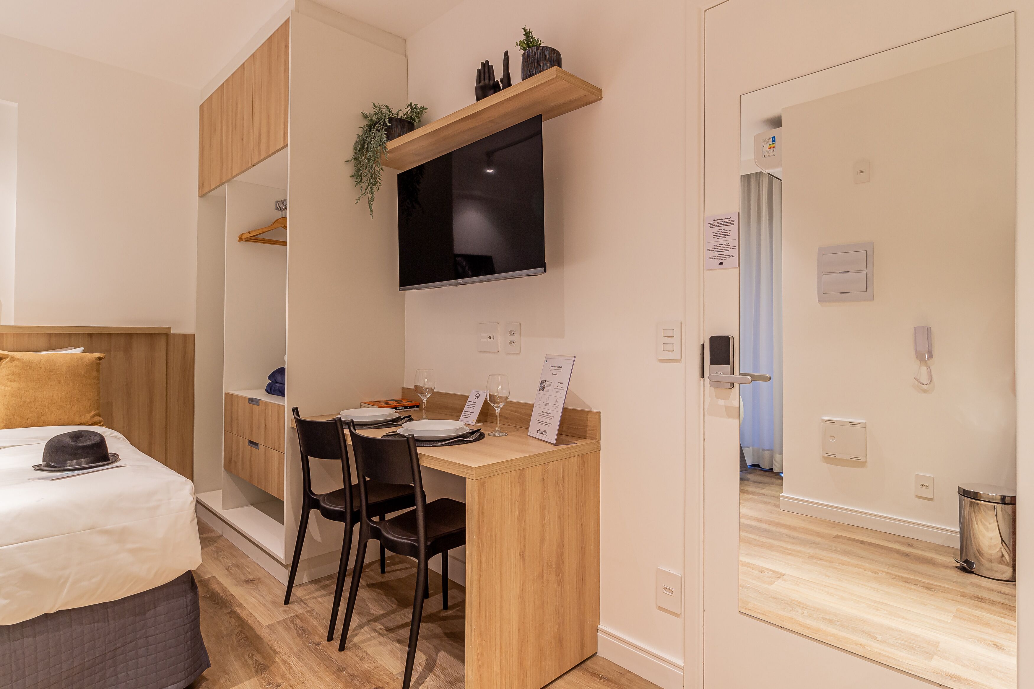 Een compacte hotelkamer met een bed, een bureau met stoel, een aan de muur bevestigde televisie en een kleine prullenbak.