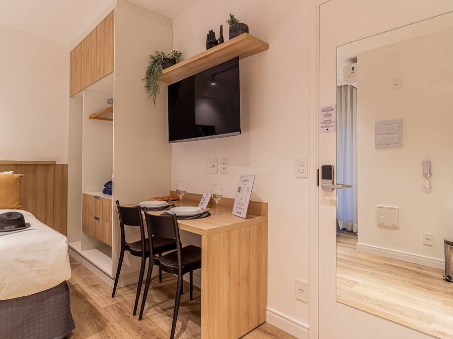 Habitación de hotel compacta con una cama, un escritorio con silla, un televisor montado en la pared y un cesto de basura pequeño.