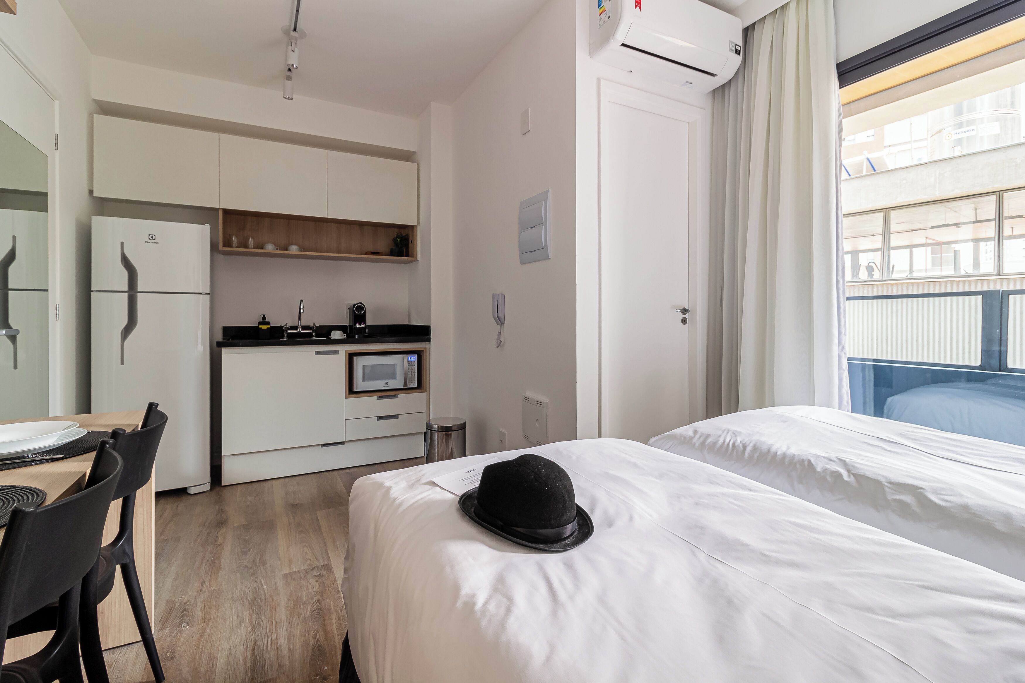 A hotel room with a bed, a dining table with chairs, a kitchenette, and a window with a view.