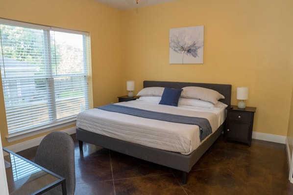 A bedroom with a bed, two bedside tables, a chair, a window with blinds, and a wall-mounted artwork.
