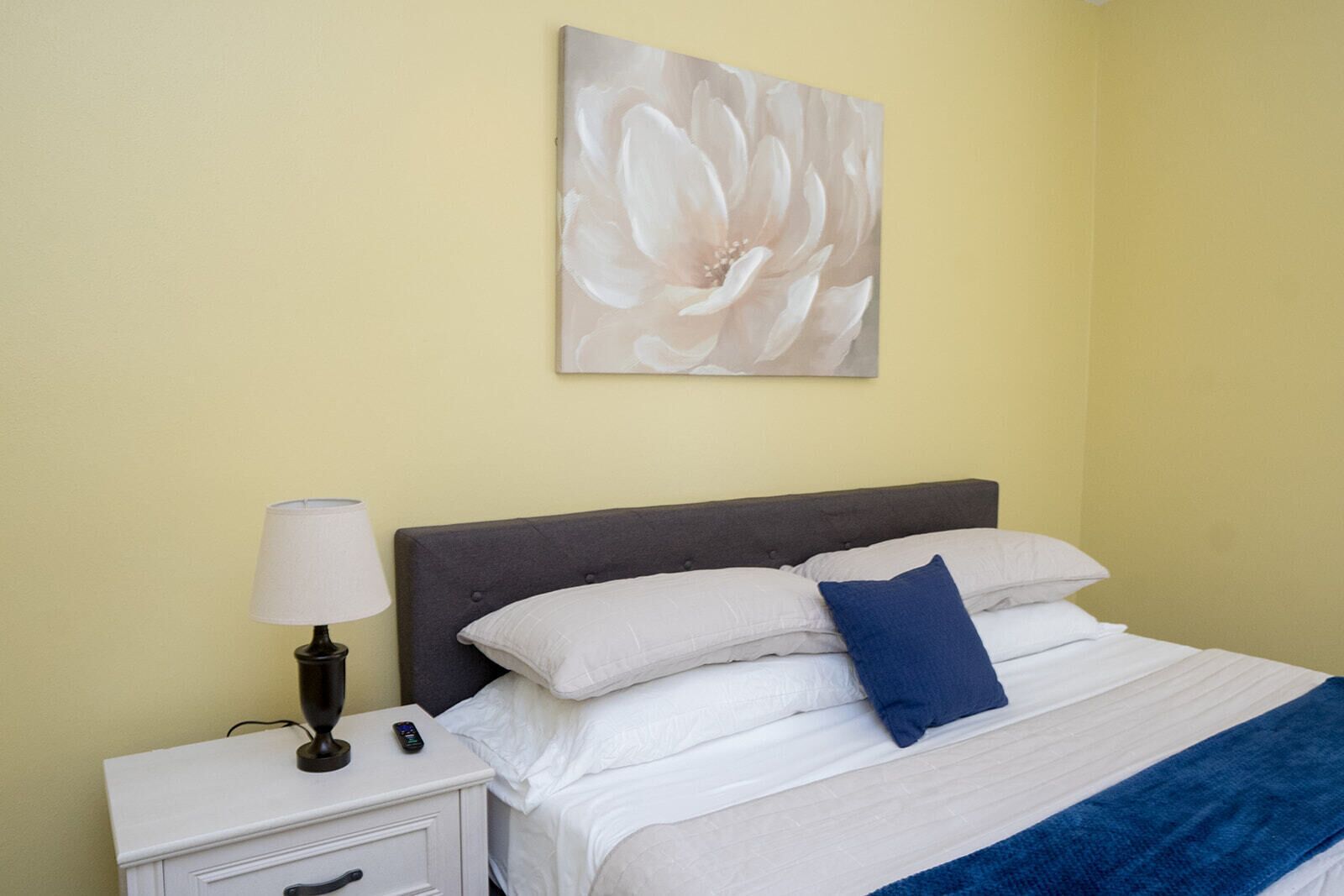 A bedroom with a bed, white bedding, a blue pillow, a nightstand with a lamp, and a wall-mounted painting.