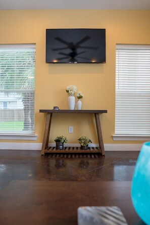 A living room with a mounted TV, a wooden console table with decorative items, and a large window with blinds. - Unit E (Moss Point)