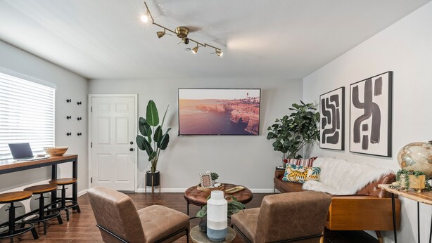 A modern living room with a brown leather sofa, a coffee table, and a TV mounted on the wall. There are potted plants and a globe on a side table. - Tribune at Redlands, Arcade Hands (Redlands)