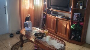 Una sala de estar con un mueble bajo de madera para el televisor, una mesa con mantel decorativo, un ventilador y una vela.