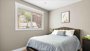 Ein Schlafzimmer mit Bett, Fenster mit Jalousien, Nachttisch mit Lampe und einem gerahmten Bild an der Wand.