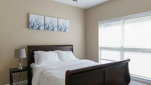 A bedroom with a wooden bed, white bedding, a nightstand with a lamp, and three abstract wall art pieces. - Unit F (Moss Point)