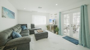 Un salon moderne avec un canapé gris, un pouf et un téléviseur à écran plat posé sur un meuble. Une porte-fenêtre coulissante donne accès à un espace extérieur aménagé avec des chaises.