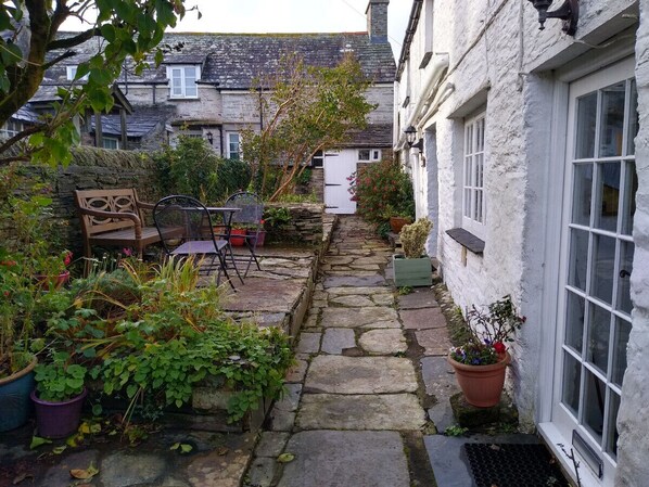 A narrow stone pathway leading to a white building with a window and a door. There are potted plants and a chair along the path. - Lilac Cottage Tintagel (Tintagel)