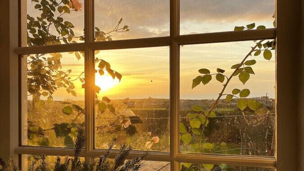 Een raam met uitzicht op een zonsondergang boven een landschap, inclusief een waterpartij en verre gebouwen.