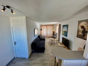 A living room with a sofa, a dining table, and a TV. There is a door on the left and a window with curtains.