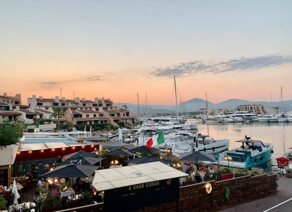 A marina with boats docked, a waterfront restaurant with outdoor seating, and a backdrop of residential buildings.