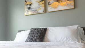 A neatly made bed with white linens and a gray throw blanket, two abstract paintings on the wall, and a light gray wall. - Unit G (Moss Point)