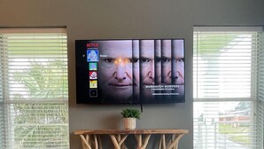 A living room with a dark wooden coffee table, a mounted flat-screen TV, and a window with blinds. - Unit G (Moss Point)