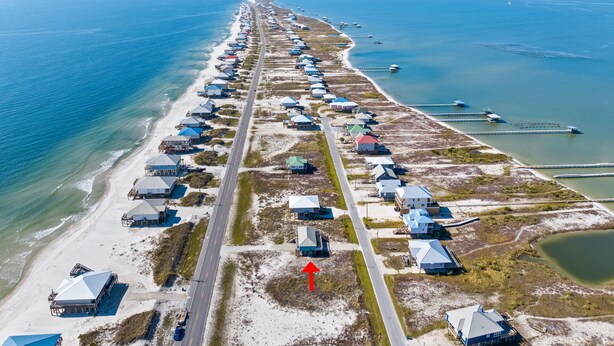 An aerial view of a coastal area with a long row of beach houses along the shoreline, a road running parallel to the beach, and a pier extending into the water.