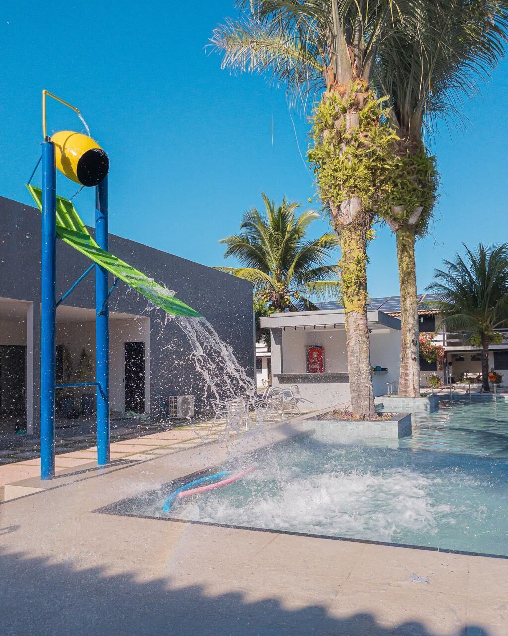 Un espace de jeux aquatiques avec un toboggan et des jets d’eau, une piscine, des palmiers et un bâtiment moderne doté d’une extincteur rouge.