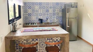 A compact kitchen with a tiled backsplash, a refrigerator, and a window with curtains. - Casa Privativa com Piscina na Ferrugem (Garopaba)