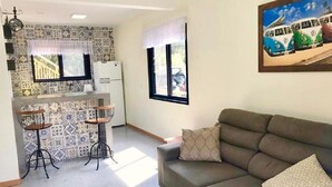 A living room with a sofa and two bar stools, a kitchen area with a refrigerator, and a patterned backsplash. - Casa Privativa com Piscina na Ferrugem (Garopaba)