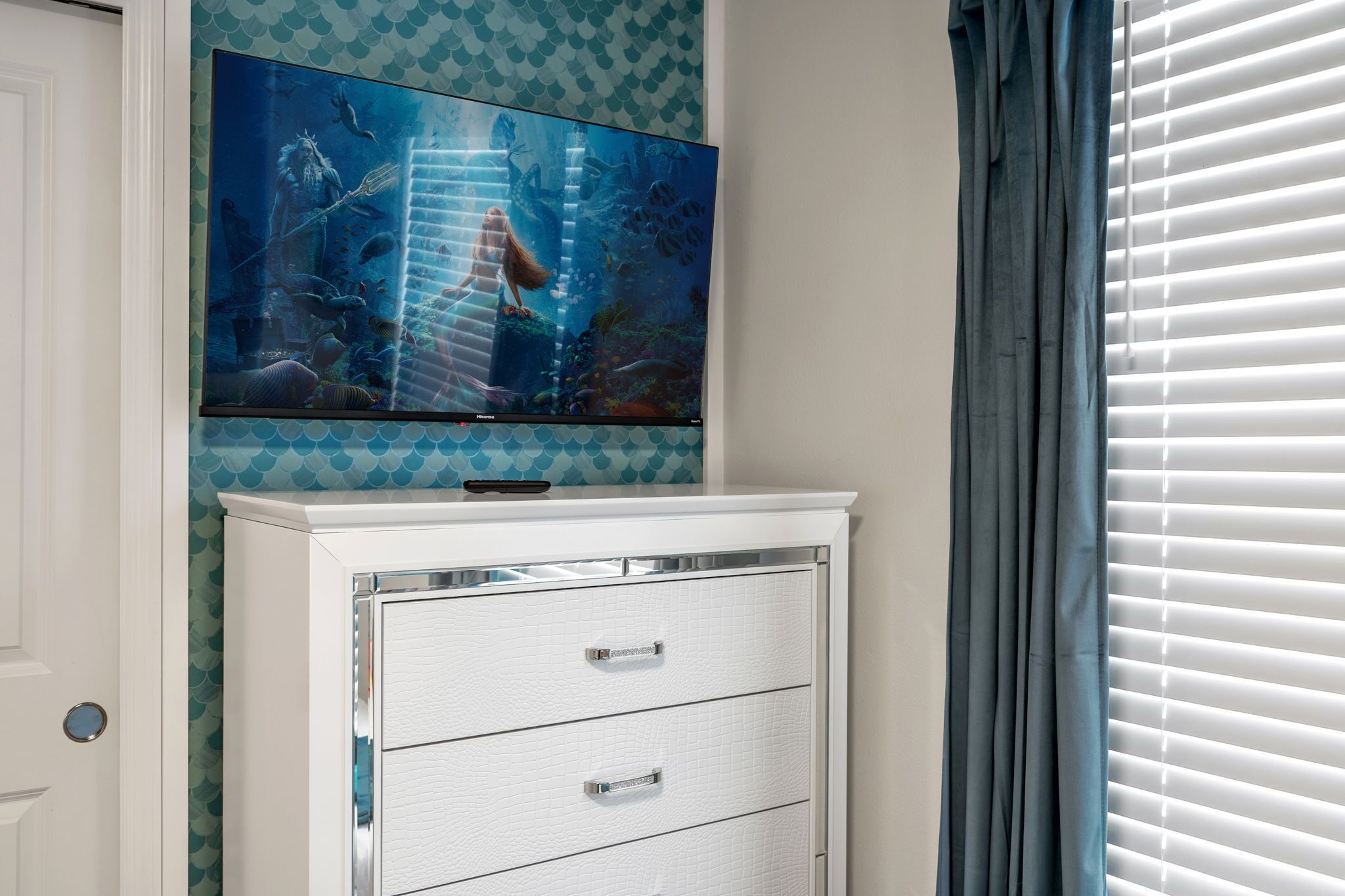 Una habitación con una televisión de pantalla plana grande montada en la pared, un aparador blanco con cajones y una ventana con persianas.