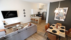 Un espacio habitable moderno con sofá gris, mesa de comedor preparada para seis personas y una cocina con isla central.