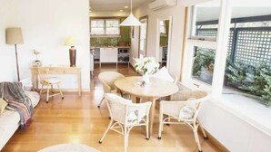 A living room with a sofa, a dining table with chairs, a kitchen in the background, and a large window with a view.