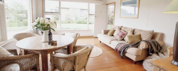 A living room with a round wooden table, wicker chairs, a sofa with cushions, and a window with a view of greenery. - Beeches By the Sea - Villa 2 (Apollo Bay)