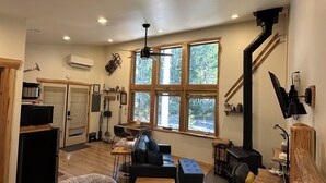 A spacious room with a large bed, a wooden desk, a black sofa, and a dining table. There are large windows providing natural light and a view of the outdoors.
