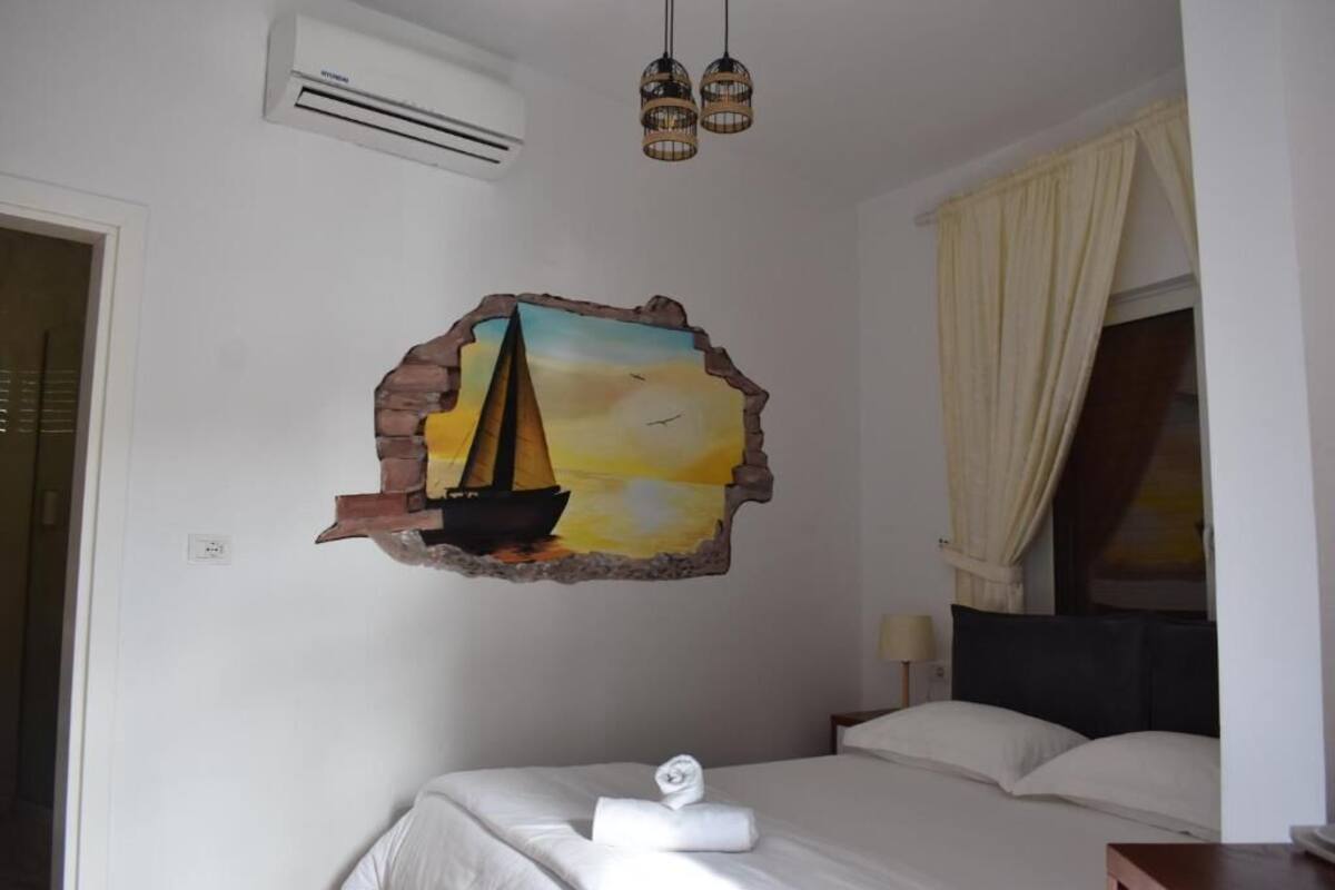 A hotel room with a bed, a wall decoration of a sailboat, an air conditioning unit, and a window with curtains.
