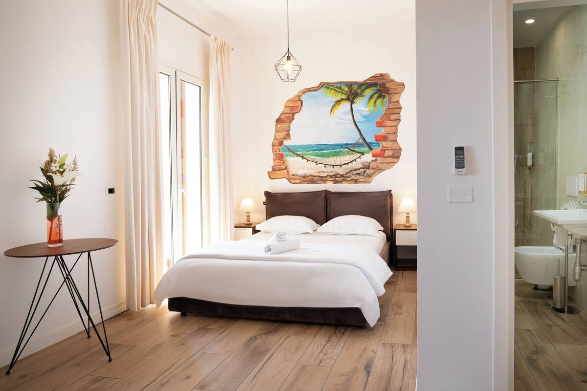 A hotel room with a bed, bedside tables, a lamp, a vase of flowers, a wall mural, a glass shower, and a bathroom with a sink and mirror.
