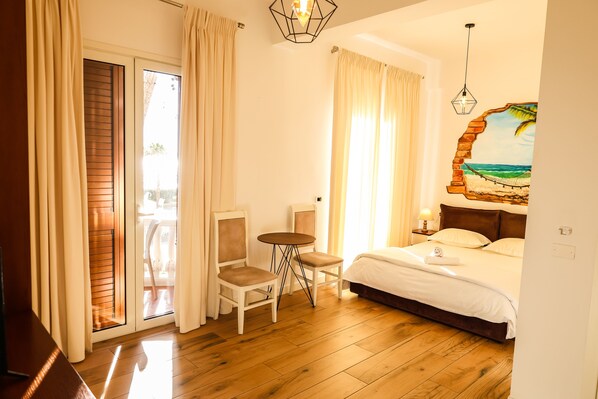 A hotel room with a bed, a small table, two chairs, a painting of a beach scene, and a balcony with curtains. - Cosmo Beach Hotel (Durrës)